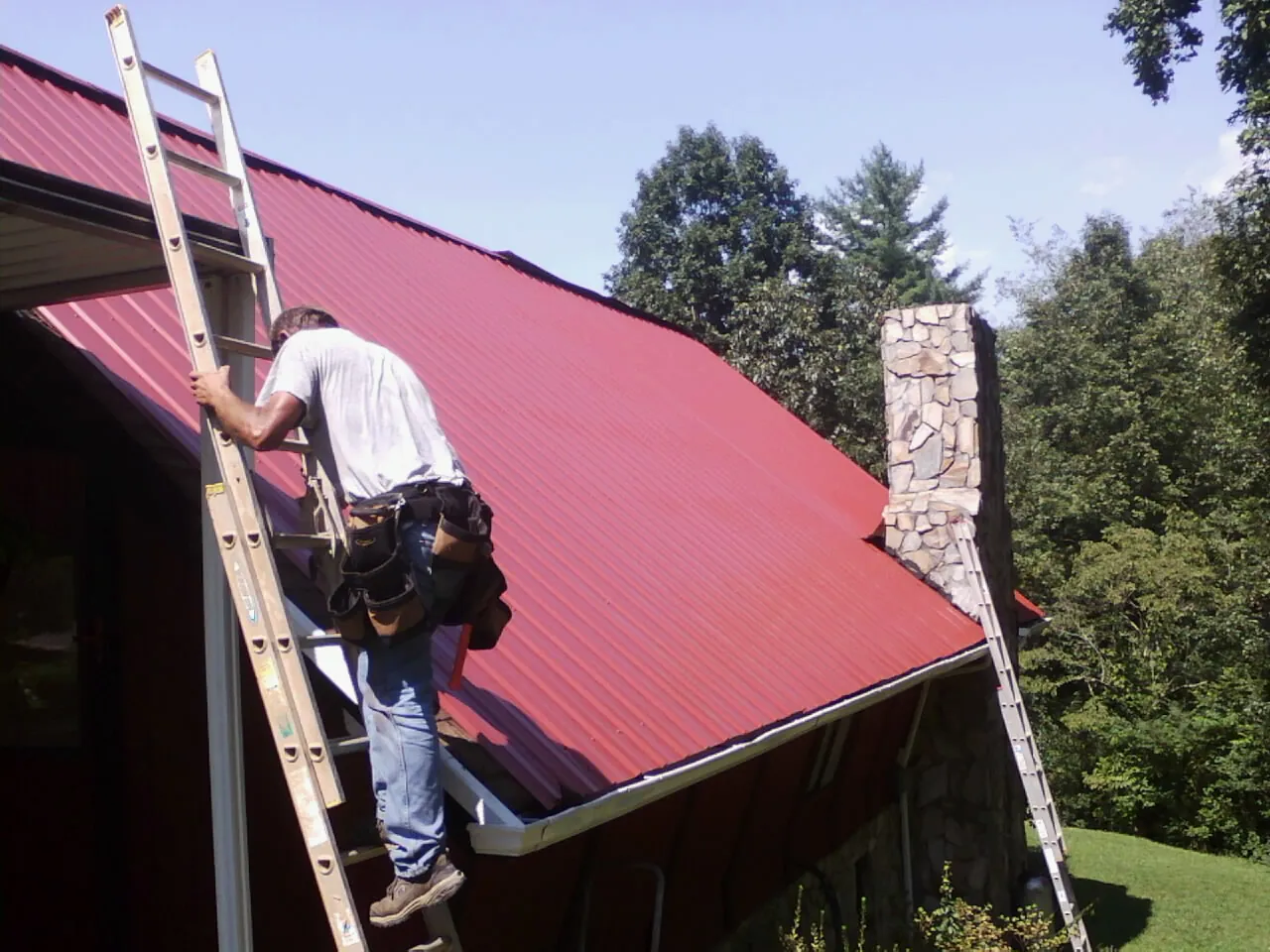 Roofer on a ladder beside a red metal roof and stone chimney — Soffit & Fascia Repair by Roofing Brothers in Pollock Pines